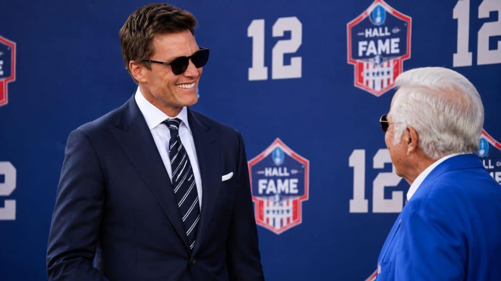 Tom Brady smiling at a Hall of Fame backdrop during a ceremonial event while interacting with an older official.