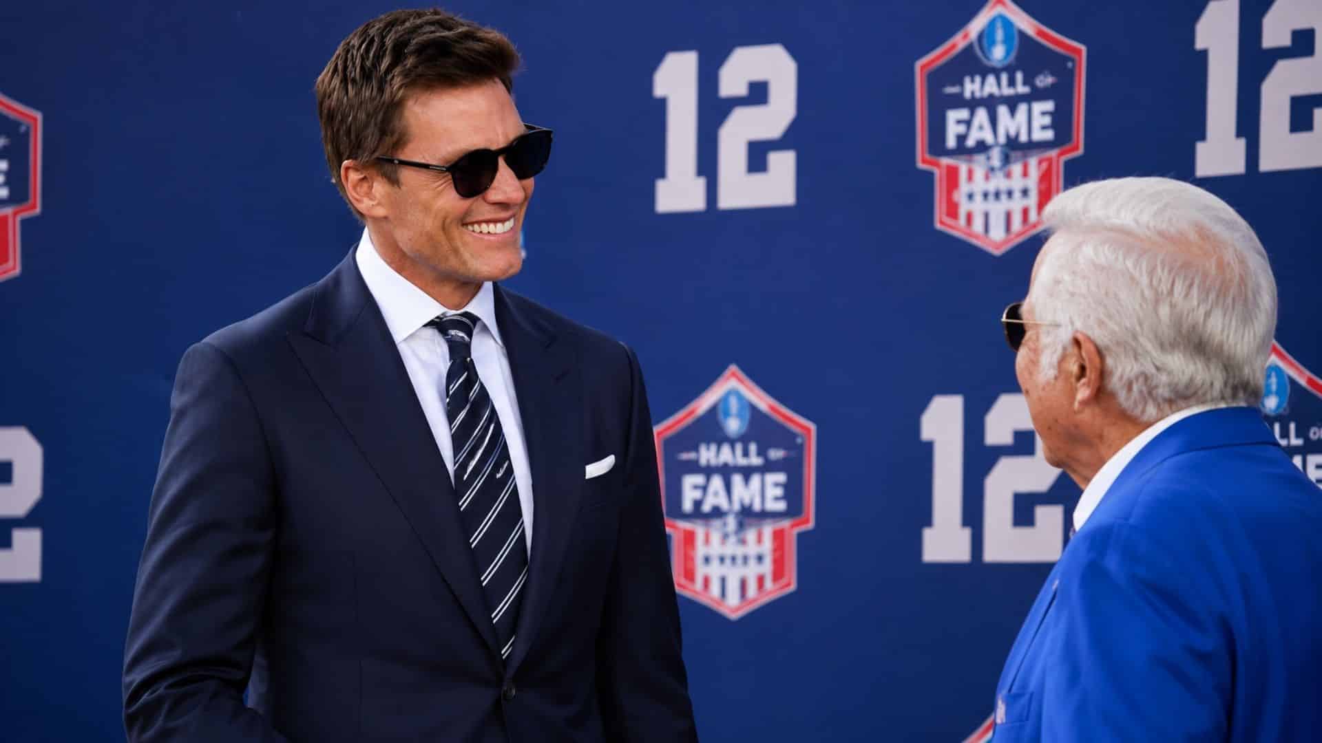 Tom Brady smiling at a Hall of Fame backdrop during a ceremonial event while interacting with an older official.