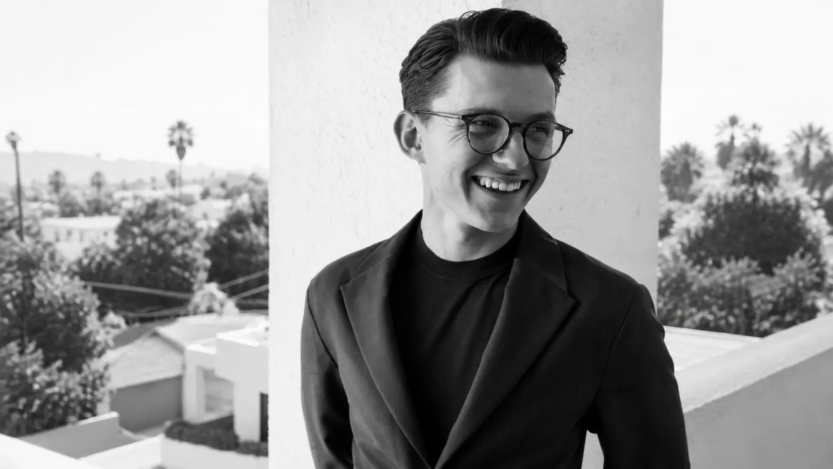 Black and white portrait of Tom Holland wearing glasses and blazer standing on balcony with blurred city view