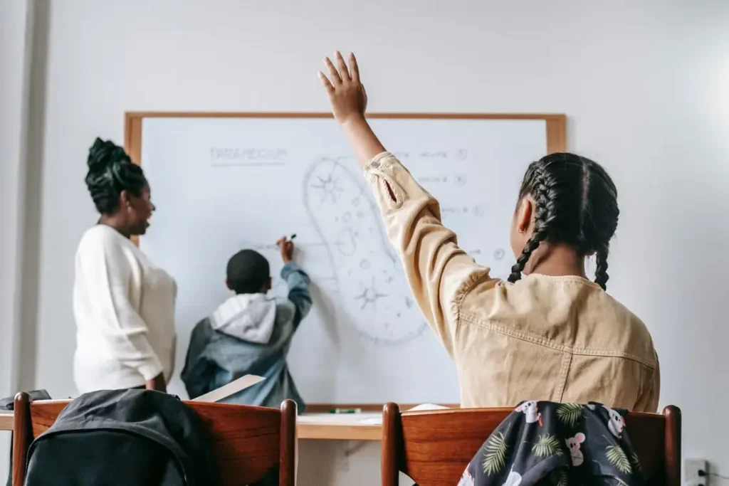 Child raising hand in classroom while teacher explains biology on whiteboard