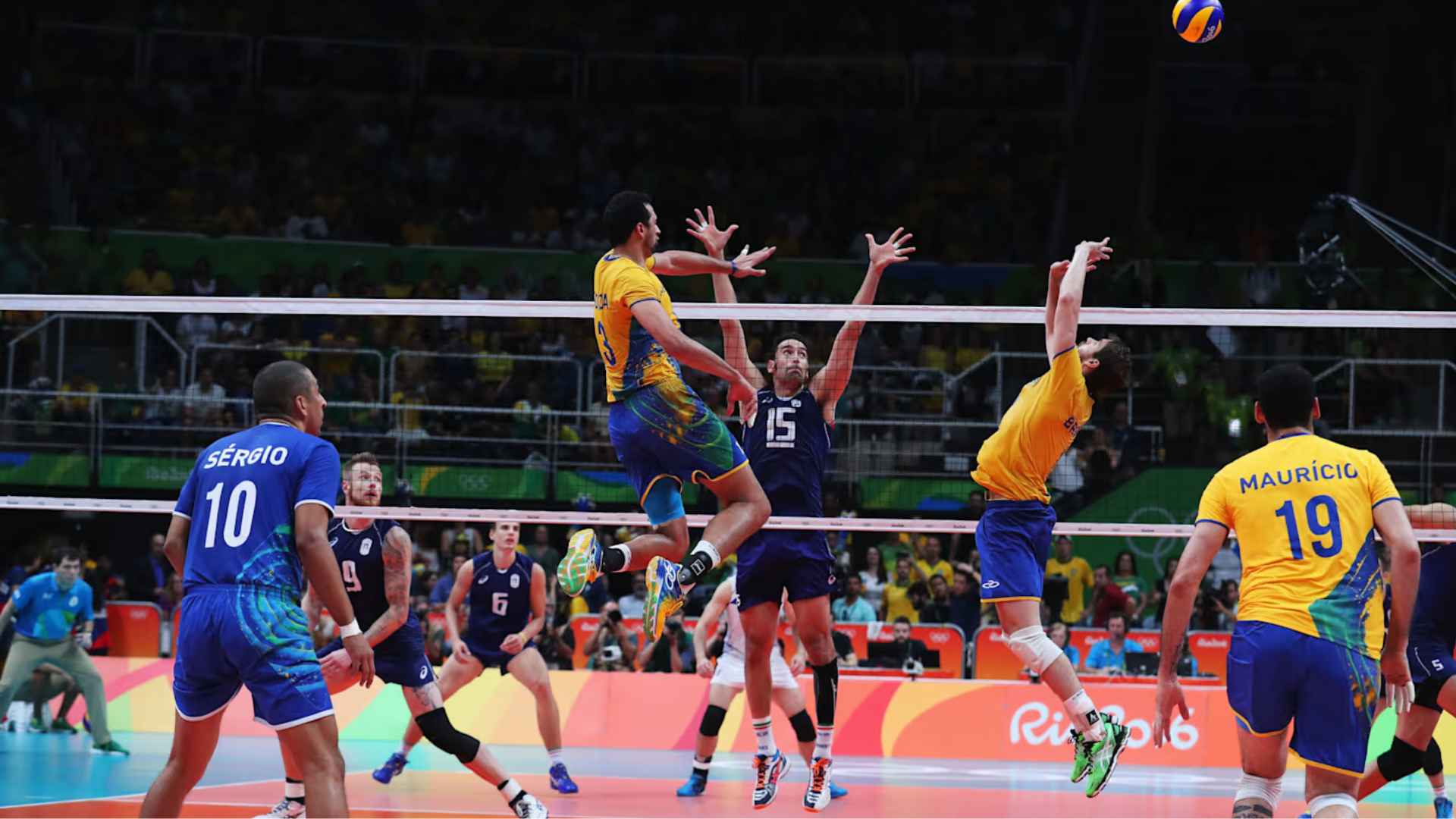 Volleyball players leap above the net during an intense international match as teammates prepare for the next play.
