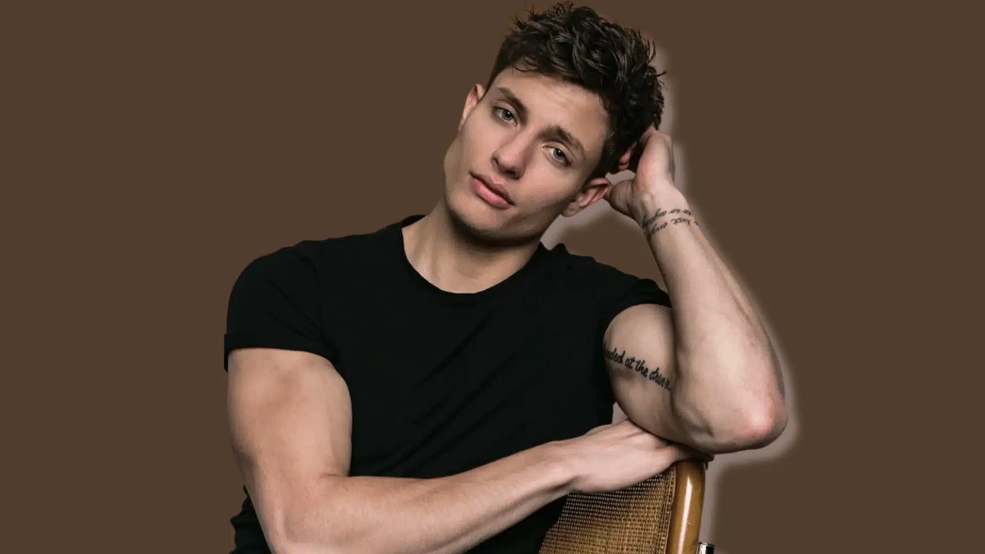 Man wearing a black shirt posing in a studio portrait with one hand resting behind his head.