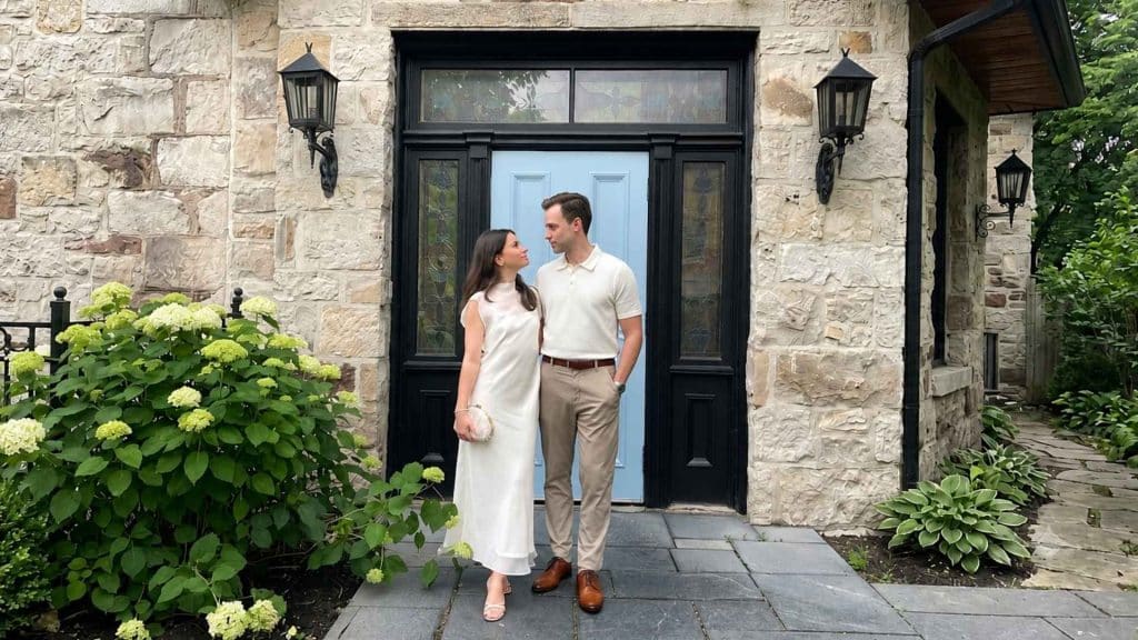Couple wearing elegant first date outfits with a white dress and beige polo standing outside a stone house