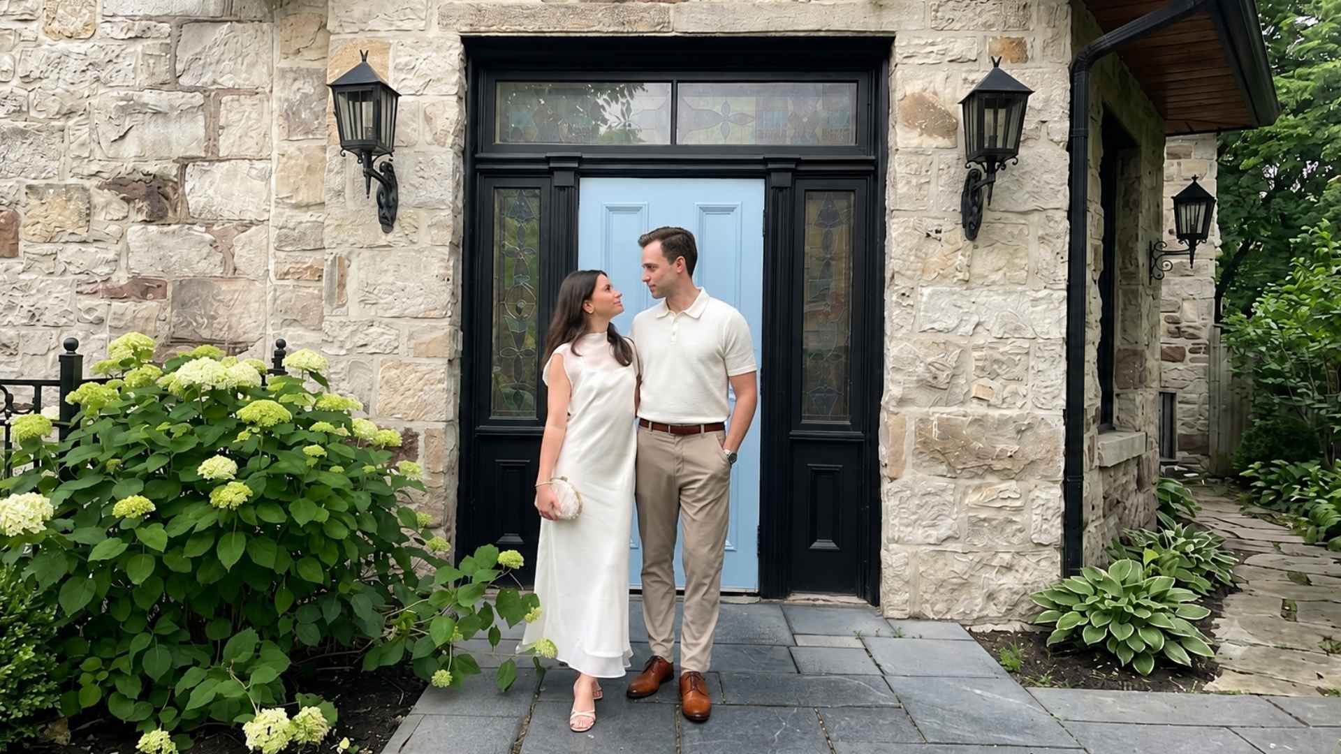 Couple wearing elegant first date outfits with a white dress and beige polo standing outside a stone house