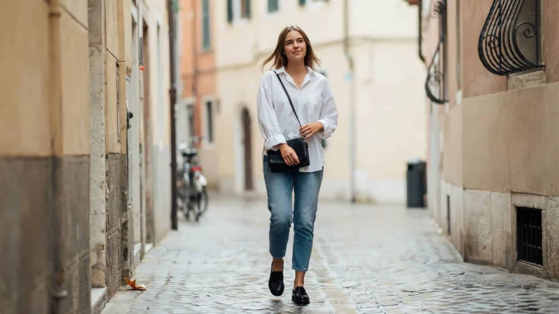 Woman wearing white button down shirt straight leg jeans and loafers walking on cobblestone street with black bag outfit