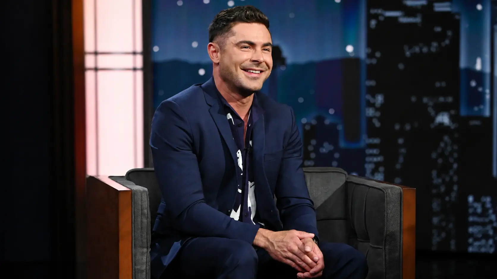 Zac efron in navy suit seated on armchair in talk show studio set with blurred city lights and blue backdrop shot
