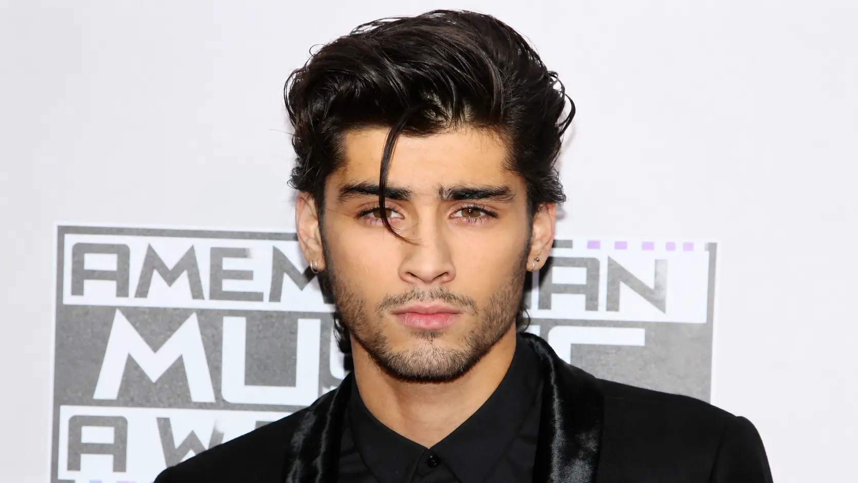 zayn malik with styled black hair wearing a black suit posing at an awards event with a white backdrop