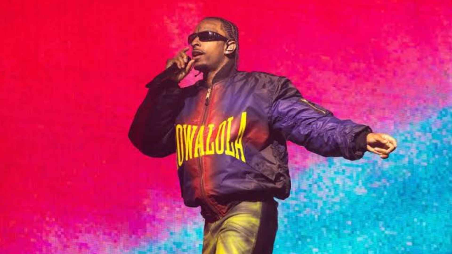21 Savage performing on stage with a microphone, wearing sunglasses and a jacket against a colorful concert background.