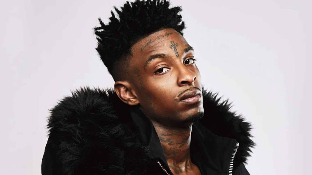 Portrait of 21 savage in a black fur-trimmed jacket, showing facial tattoos against a light background.