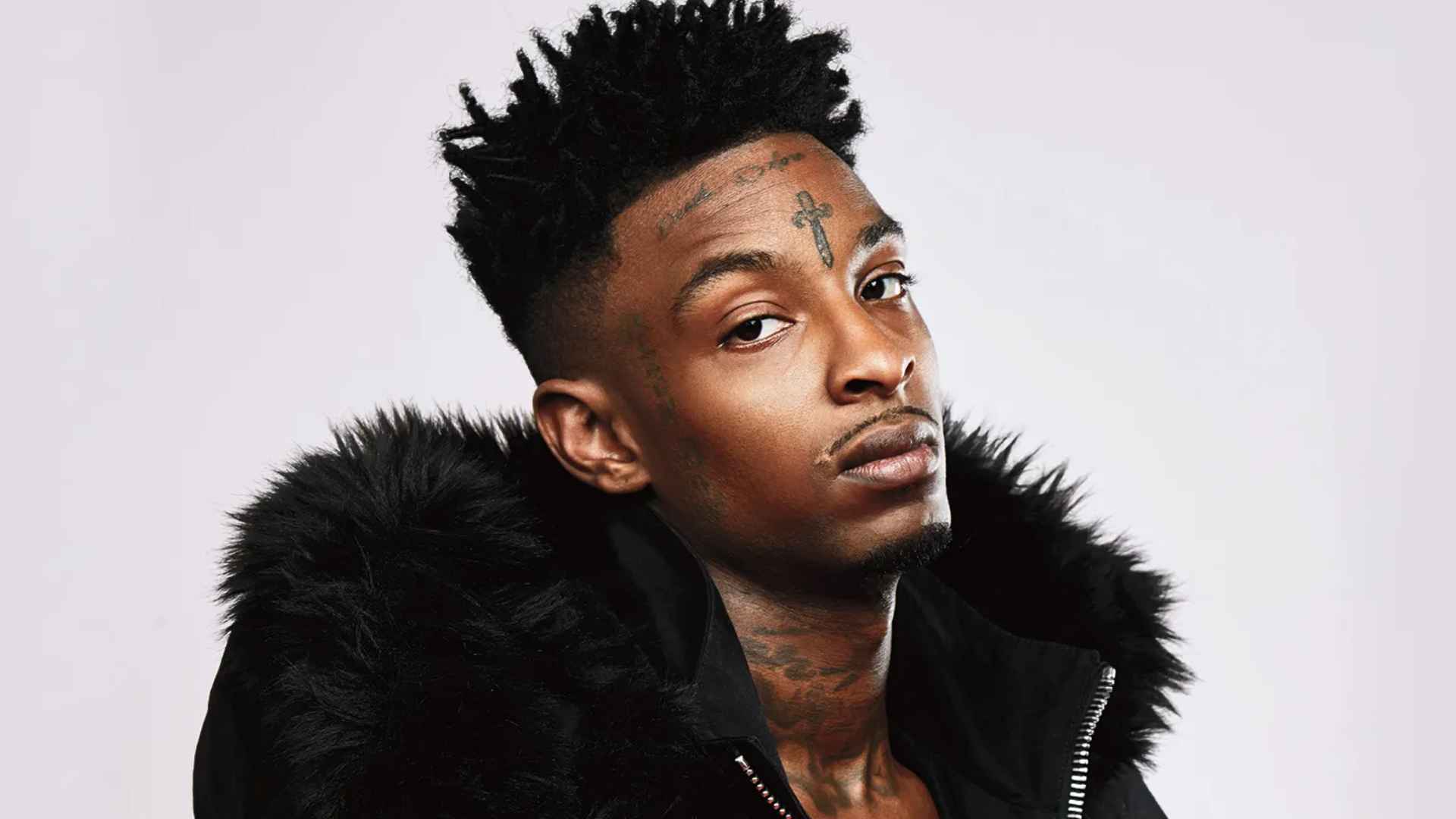 Portrait of 21 savage in a black fur-trimmed jacket, showing facial tattoos against a light background.