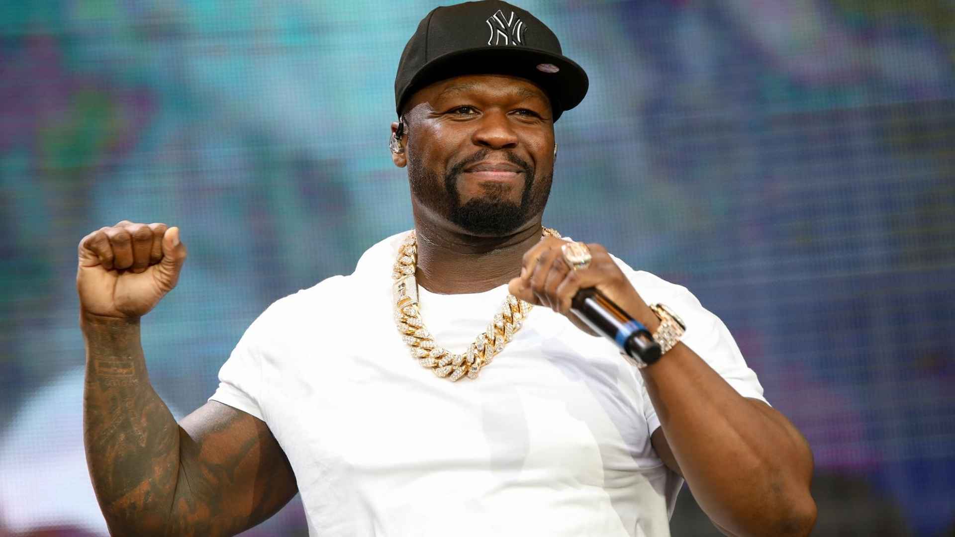 50 cents performing live on stage wearing a white shirt and cap while holding a microphone with jewelry visible