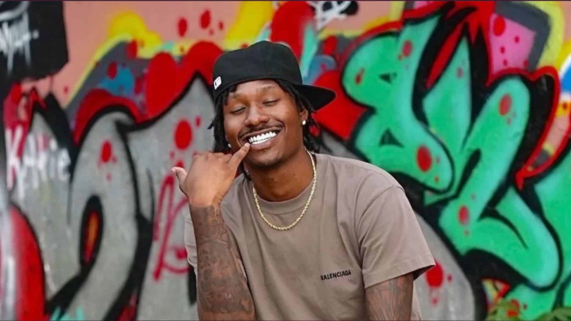 Duke Dennis wearing a black cap and beige shirt posing in front of colorful graffiti artwork background