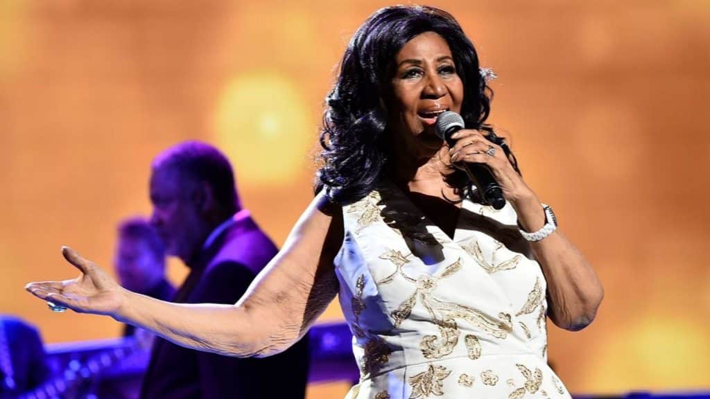 Aretha Franklin performs live onstage in a white and gold sequined dress, singing into a microphone with one arm outstretched.