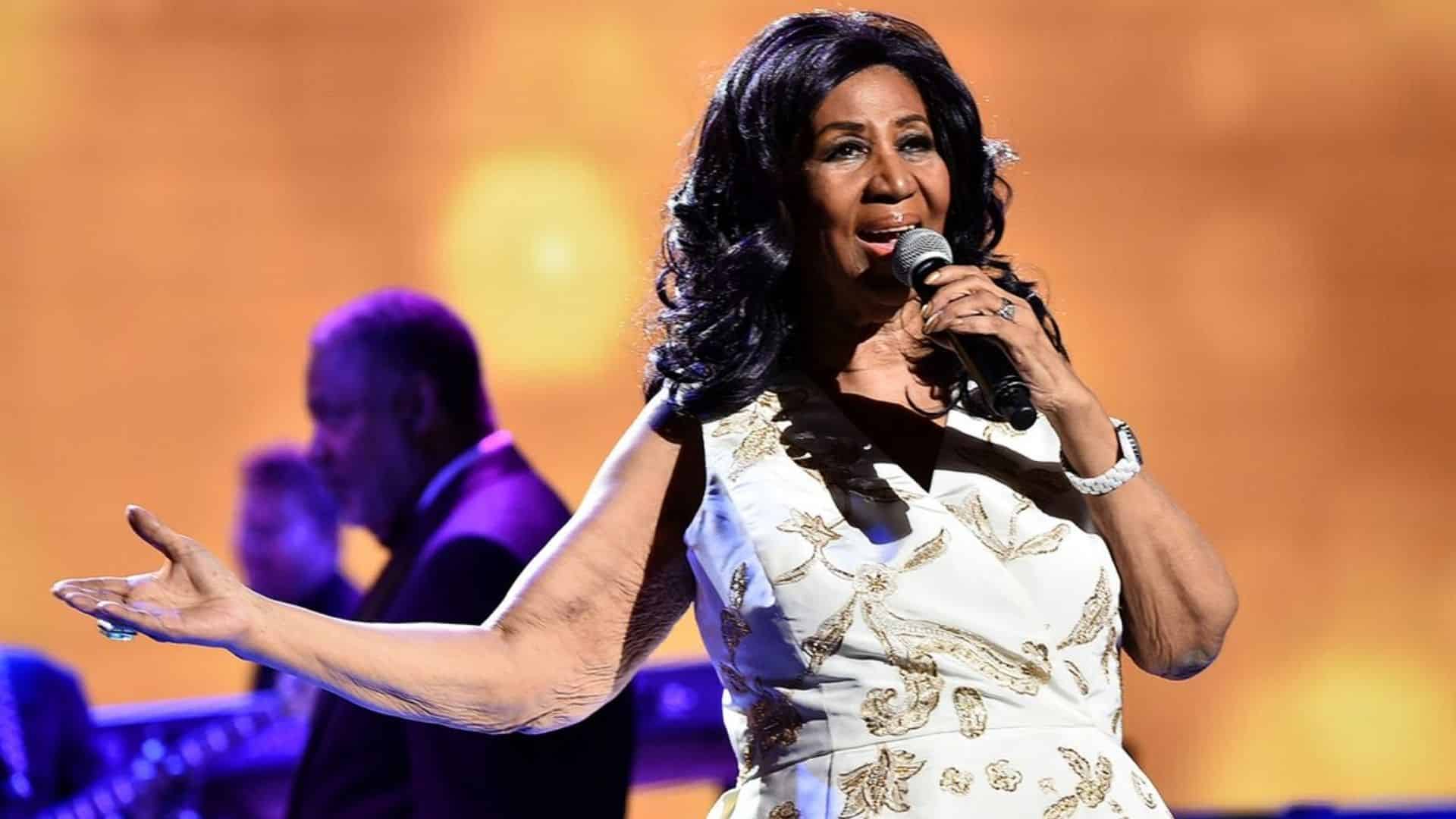Aretha Franklin performs live onstage in a white and gold sequined dress, singing into a microphone with one arm outstretched.
