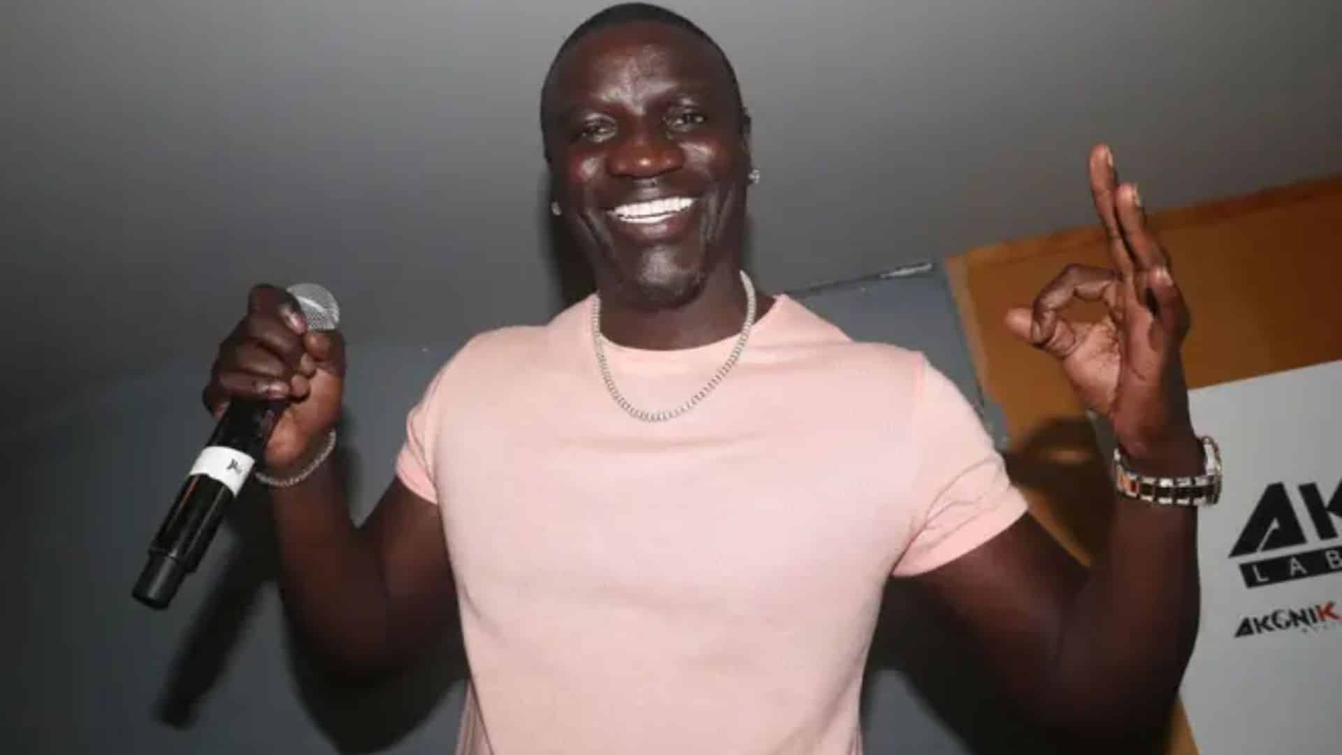 Akon performing on stage while holding a microphone and smiling during a live music event with audience atmosphere