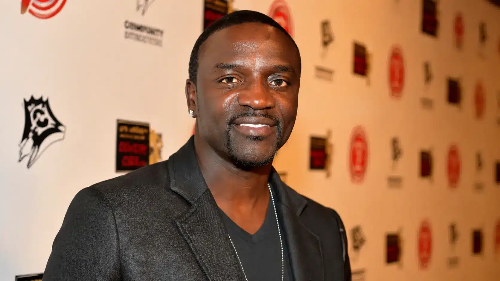 Akon posing in a black blazer with chain necklace at a media event featuring soft background logos and lights