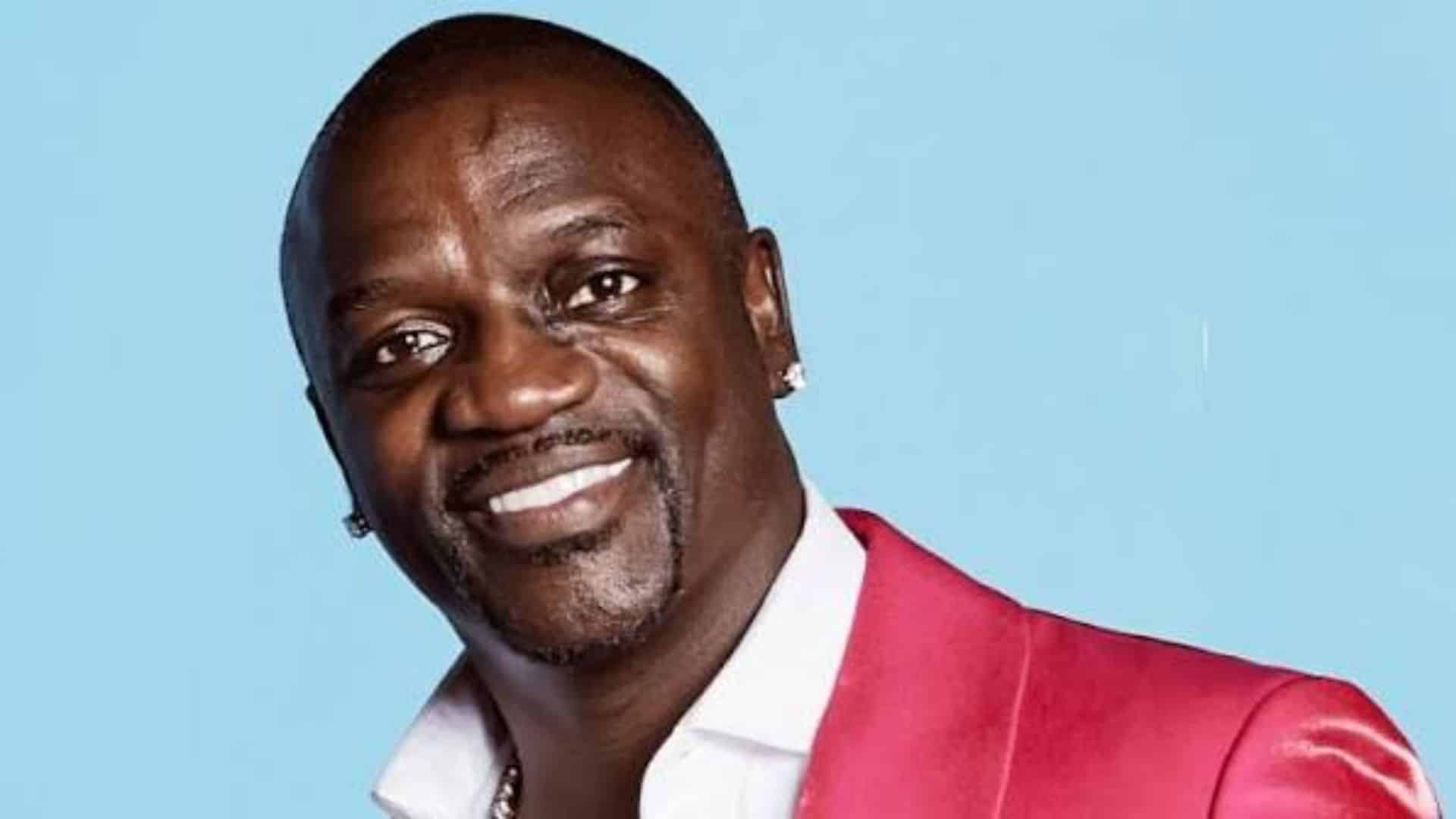 Akon posing for a portrait wearing a red jacket with a smile against a blue background during a public appearance
