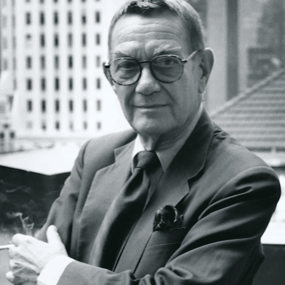 Black-and-white portrait of Albert Hadley in glasses and dark suit standing outdoors with city buildings in background.