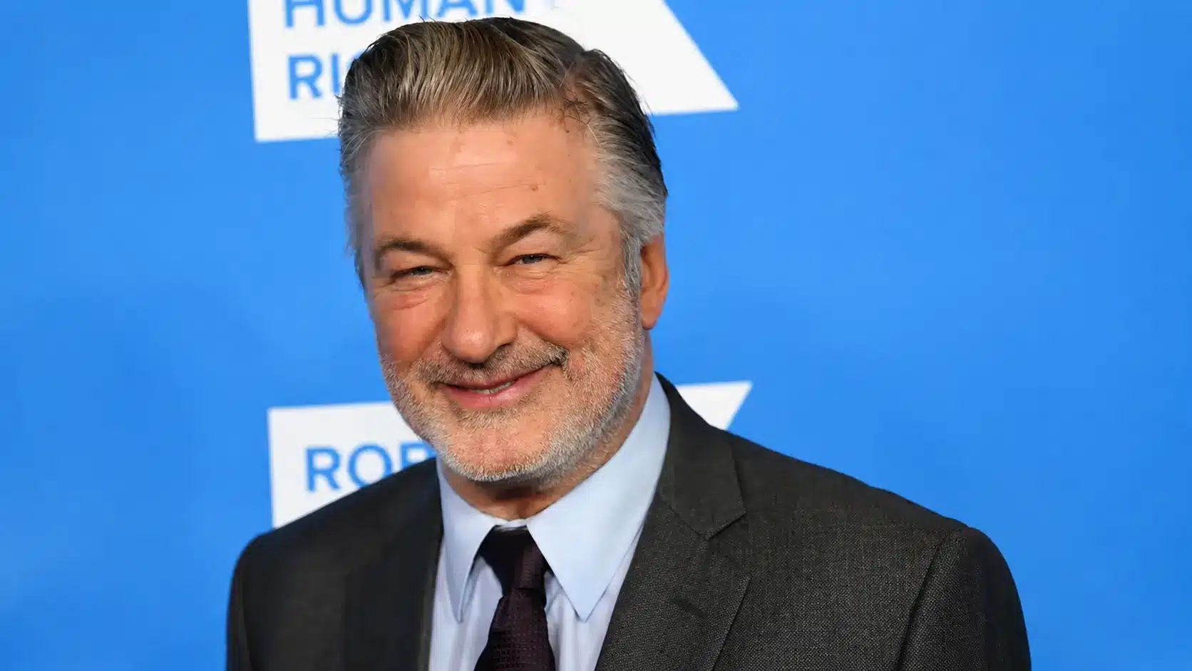 Alec Baldwin in a dark suit and tie posing against a blue event backdrop with bright lighting and formal portrait style