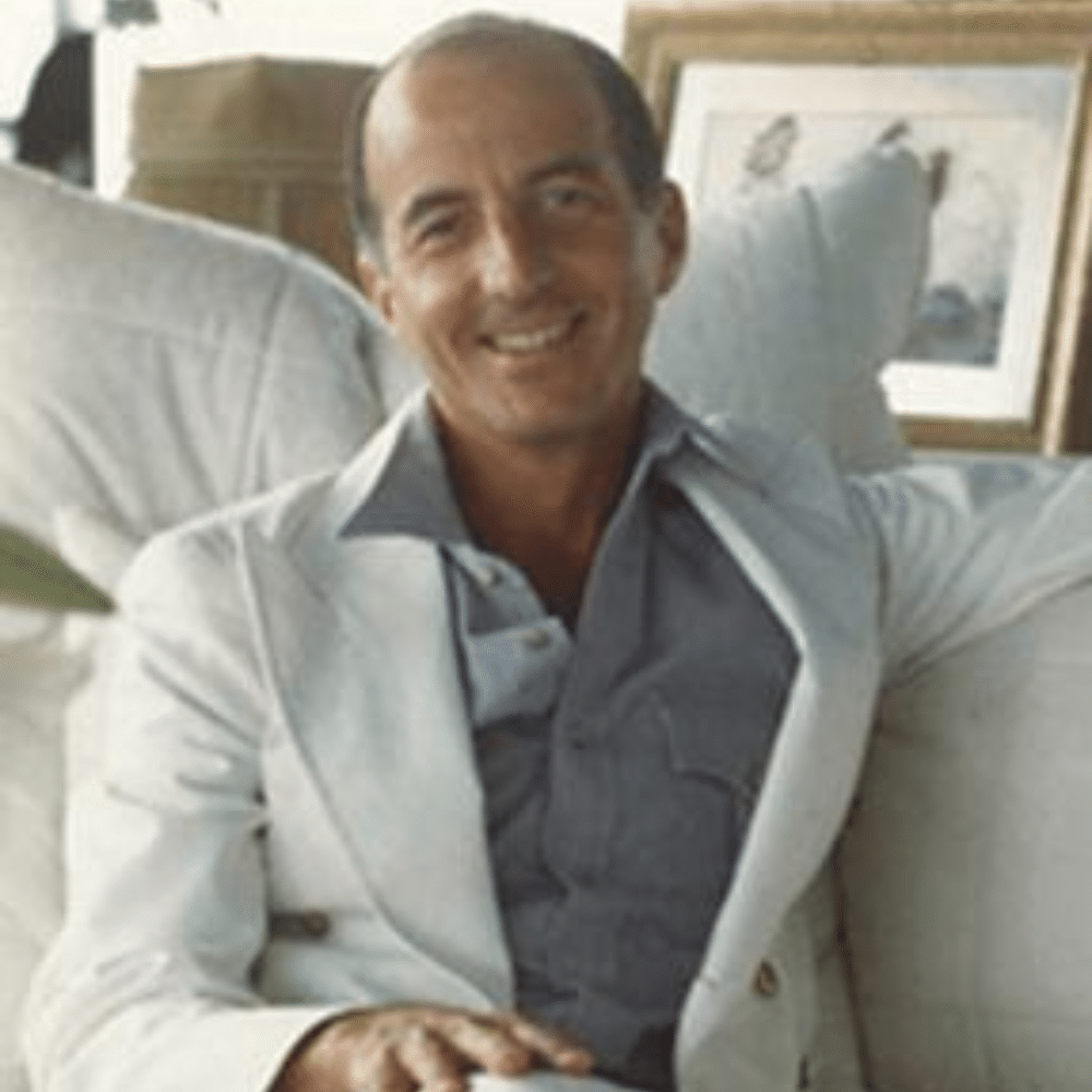 Angelo Donghia smiling in white suit seated on sofa in bright living room with framed artwork in background.