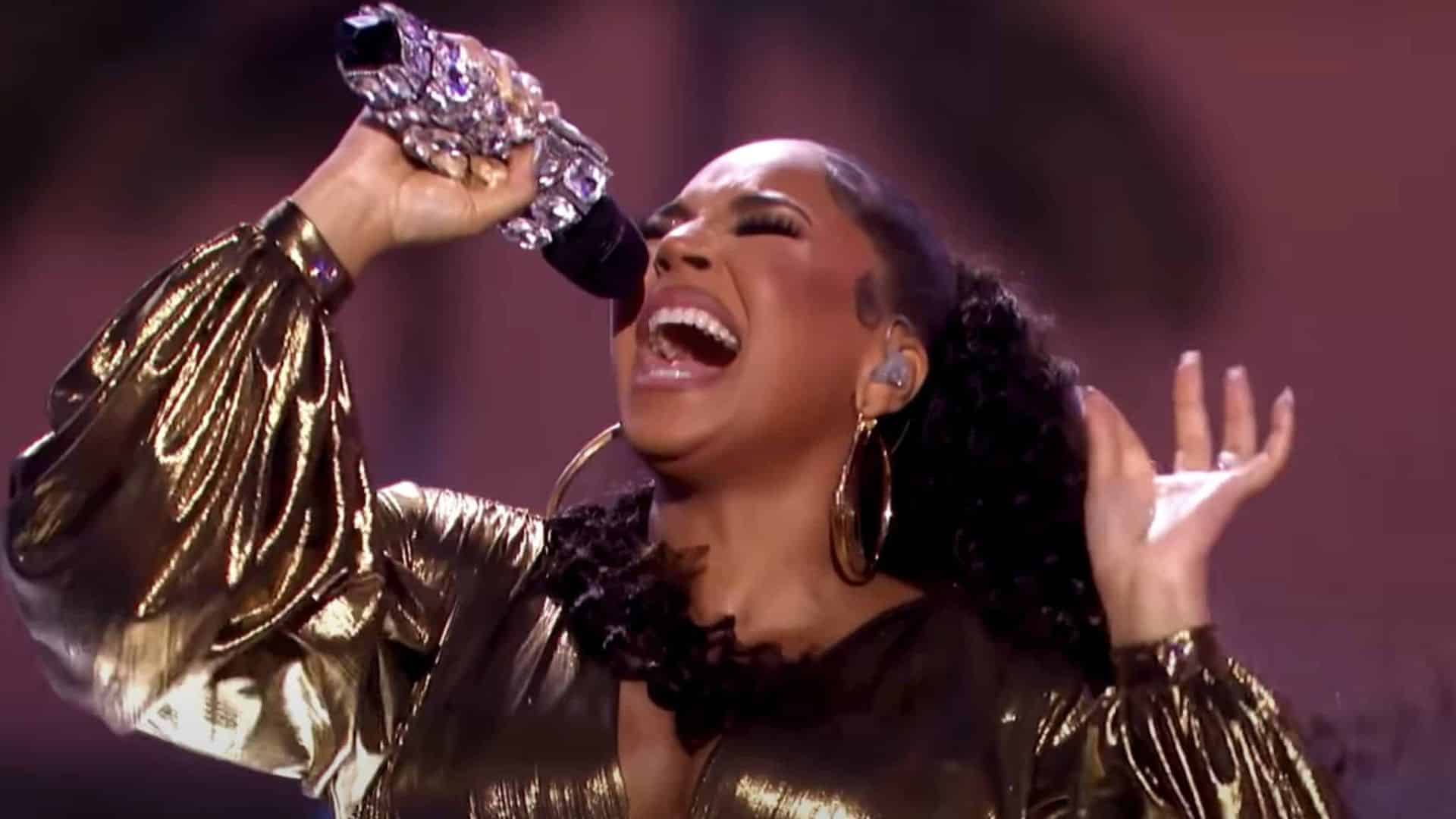 Ashanti singing into a microphone during a live performance while wearing a metallic outfit and stage lighting