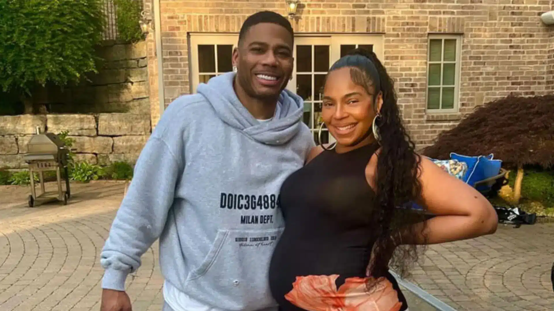 Ashanti posing with her husband outdoors in a backyard setting while wearing a black outfit with a brick house background