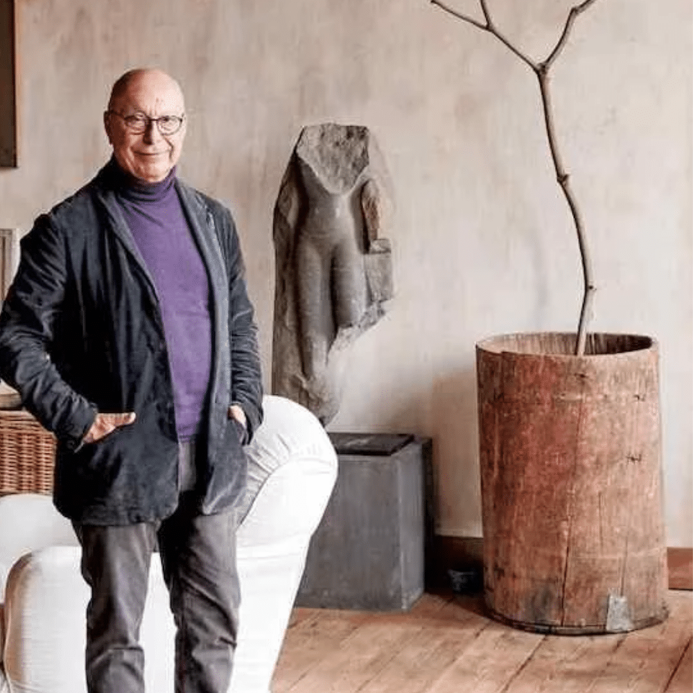 Axel Vervoordt standing beside white chair and sleeping dog in rustic room with sculpture and large potted branch.