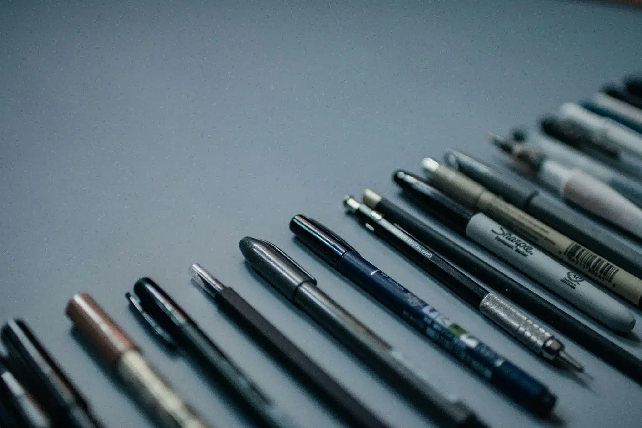 Various pens and markers arranged on a smooth gray surface in soft lighting