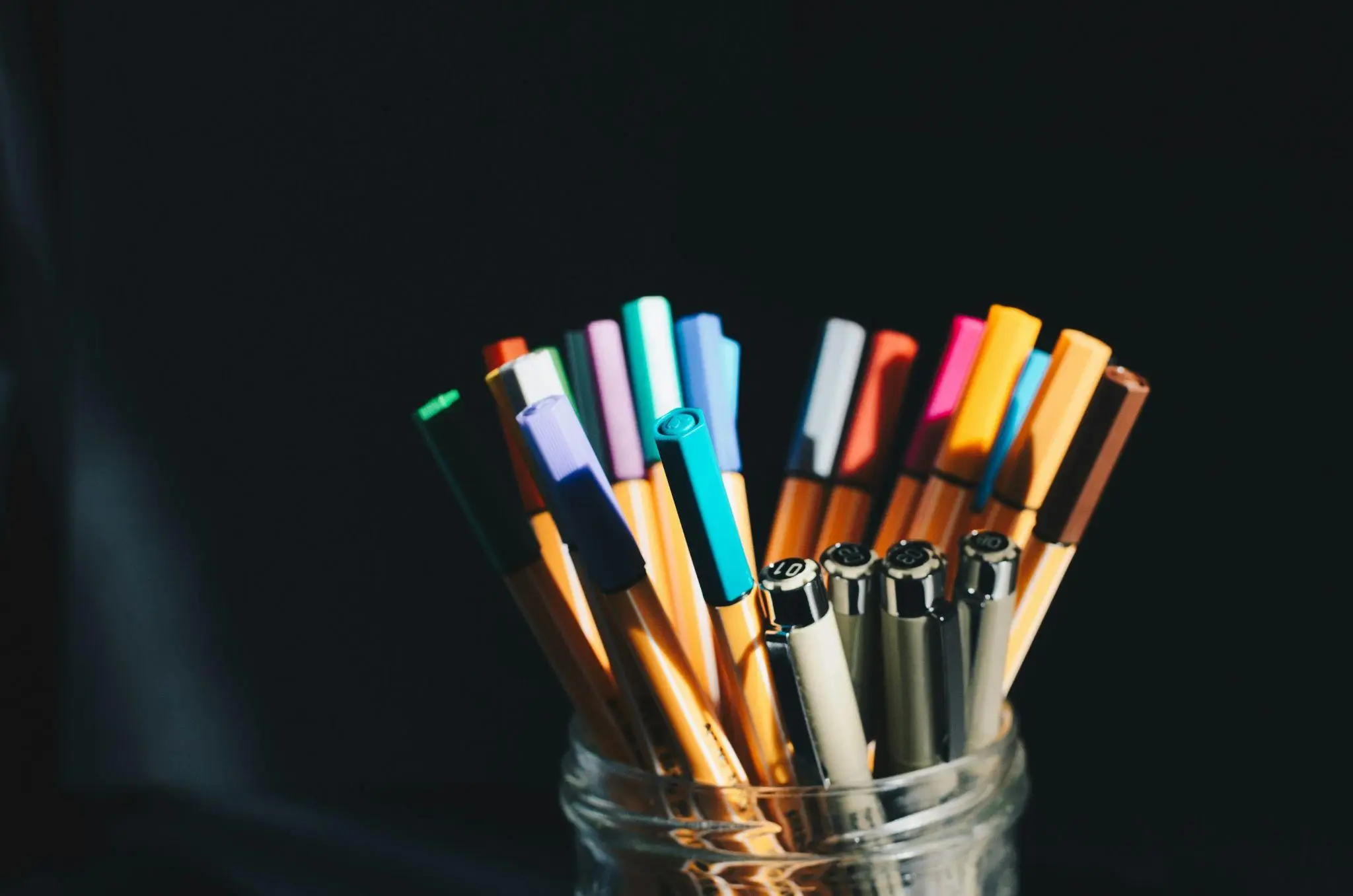 Colorful pens and markers in a glass jar against a dark background