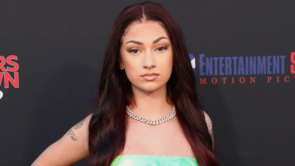 Bhad Bhabie posing at a red-carpet event in a green strapless outfit with long dark red hair and a silver chain necklace.