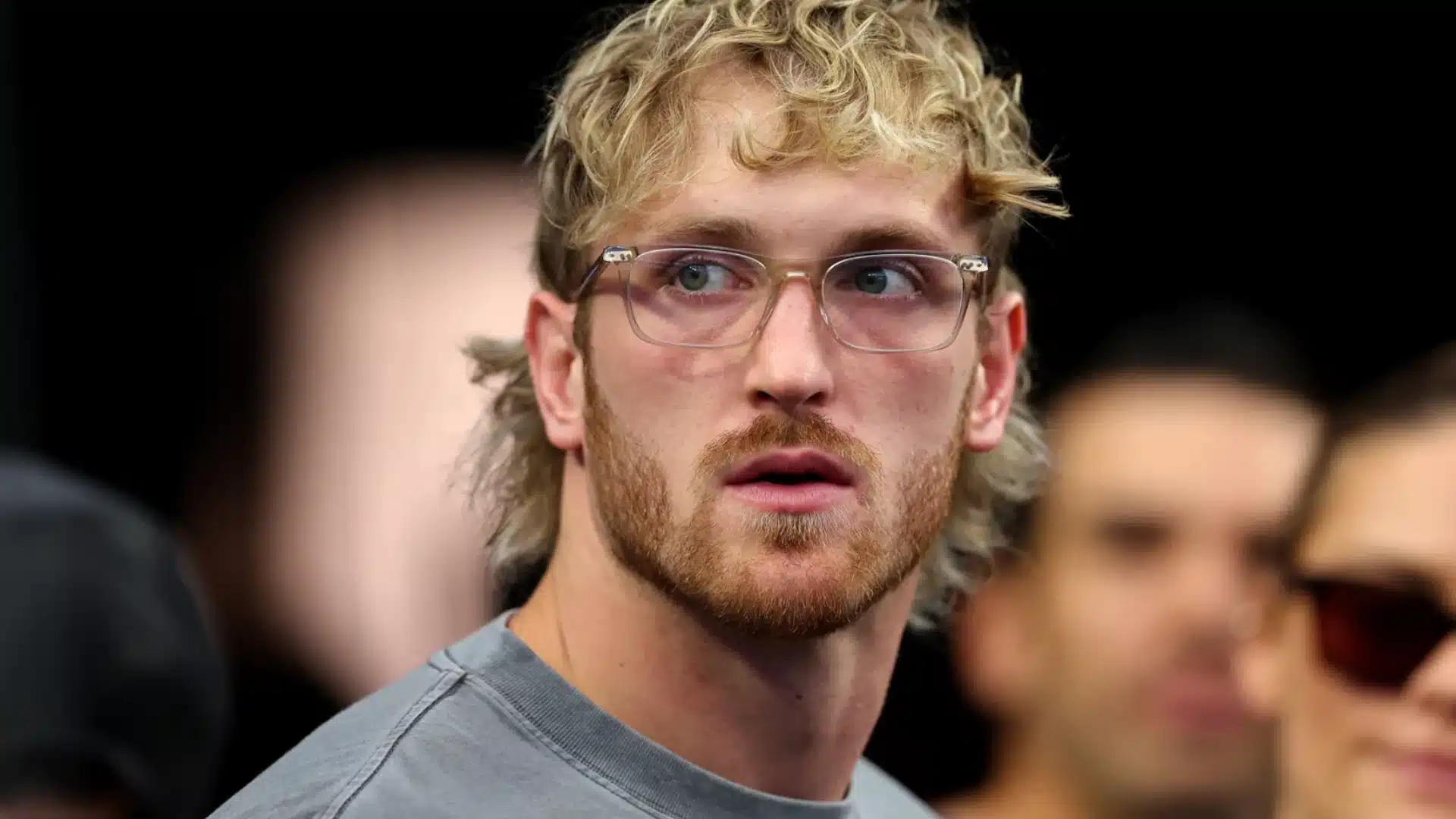 Blond male creator wearing clear glasses and a gray shirt at a crowded public event