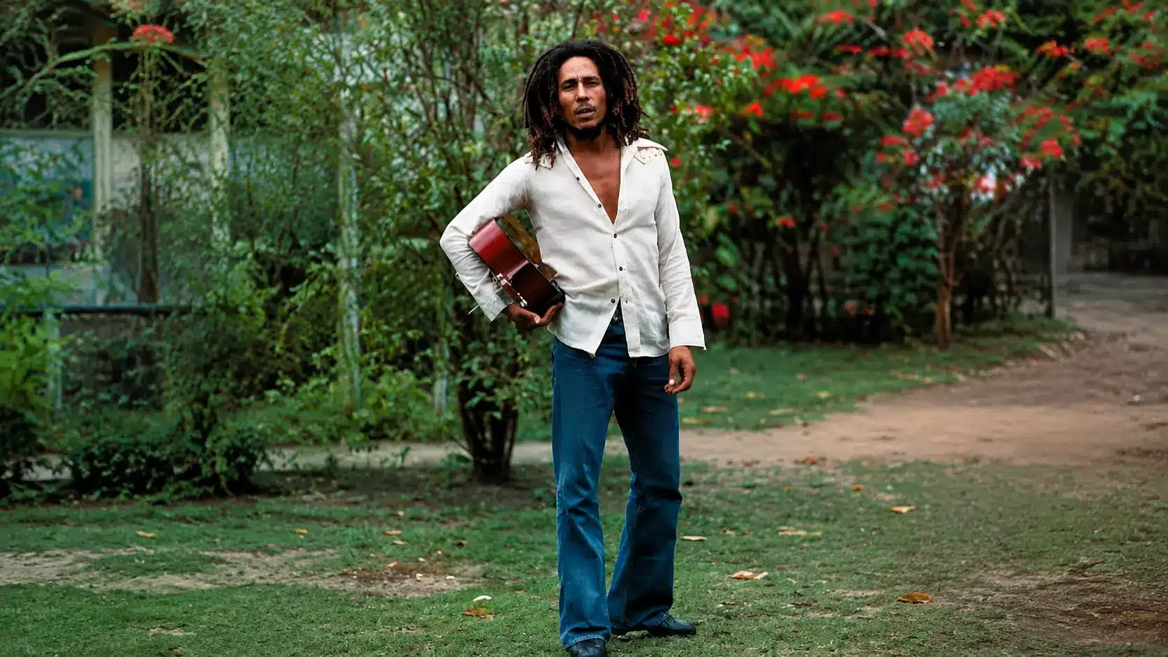 Bob Marley standing in a green garden holding a guitar with a cream shirt and blue jeans surrounded by trees and flowers