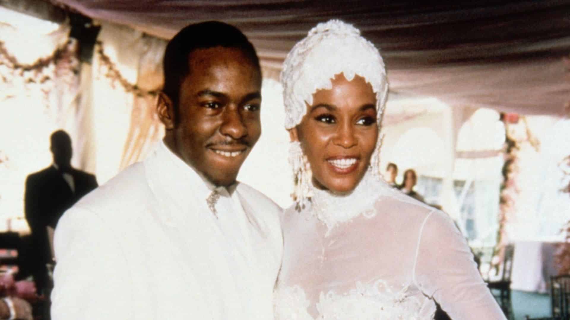 Bobby Brown standing beside Whitney Houston at a formal event while both wear white outfits and smile together
