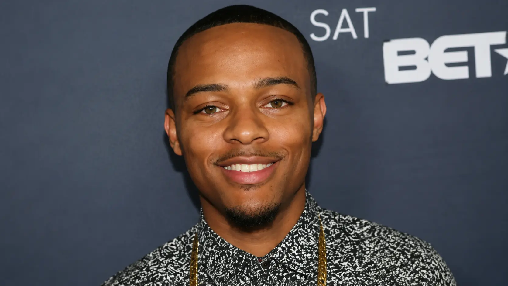 Close portrait of Bow Wow with short hair wearing printed shirt and necklace against a branded event background