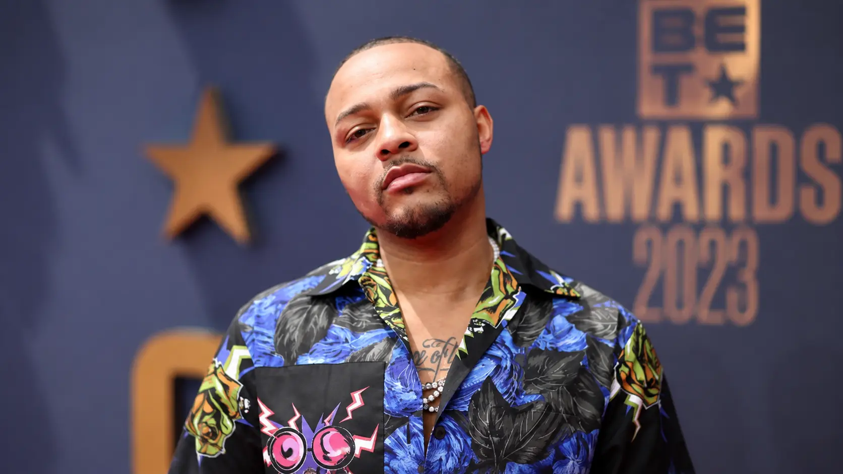 Bow Wow wearing a colorful patterned shirt at a red carpet event with blurred award backdrop and star detail behind him