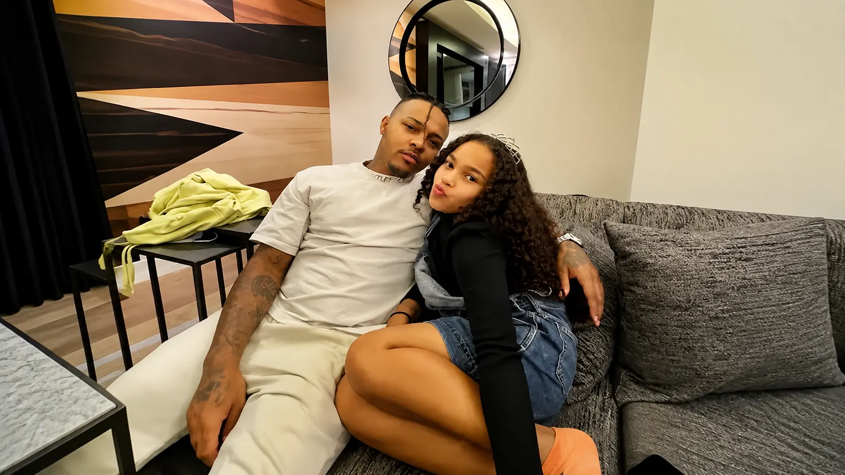 Bow wow and his daughter sitting together on a gray sofa in modern living room with wall art mirror and cozy indoor decor