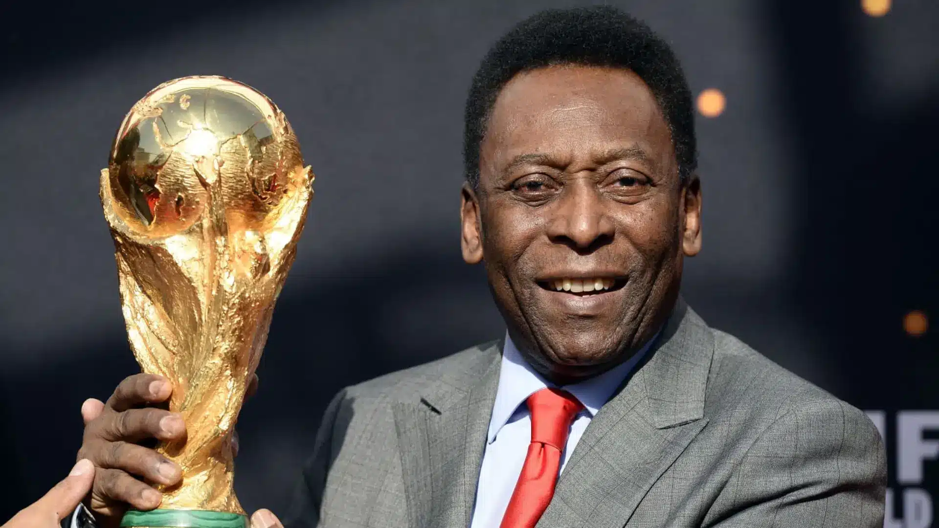 Brazilian soccer legend Pelé smiling while holding the golden FIFA World Cup trophy, wearing a suit and red tie.