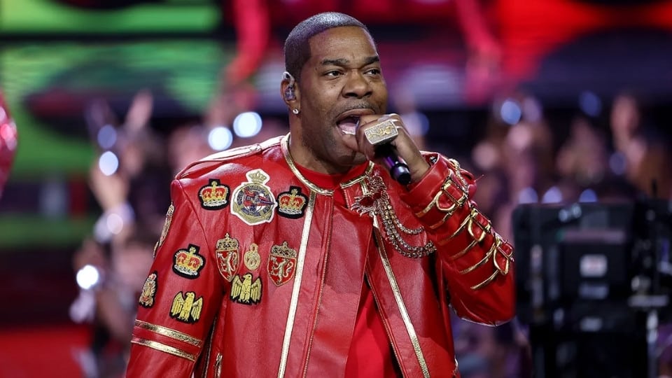 Busta Rhymes performing on stage in a vibrant red leather jacket adorned with decorative crests and patches