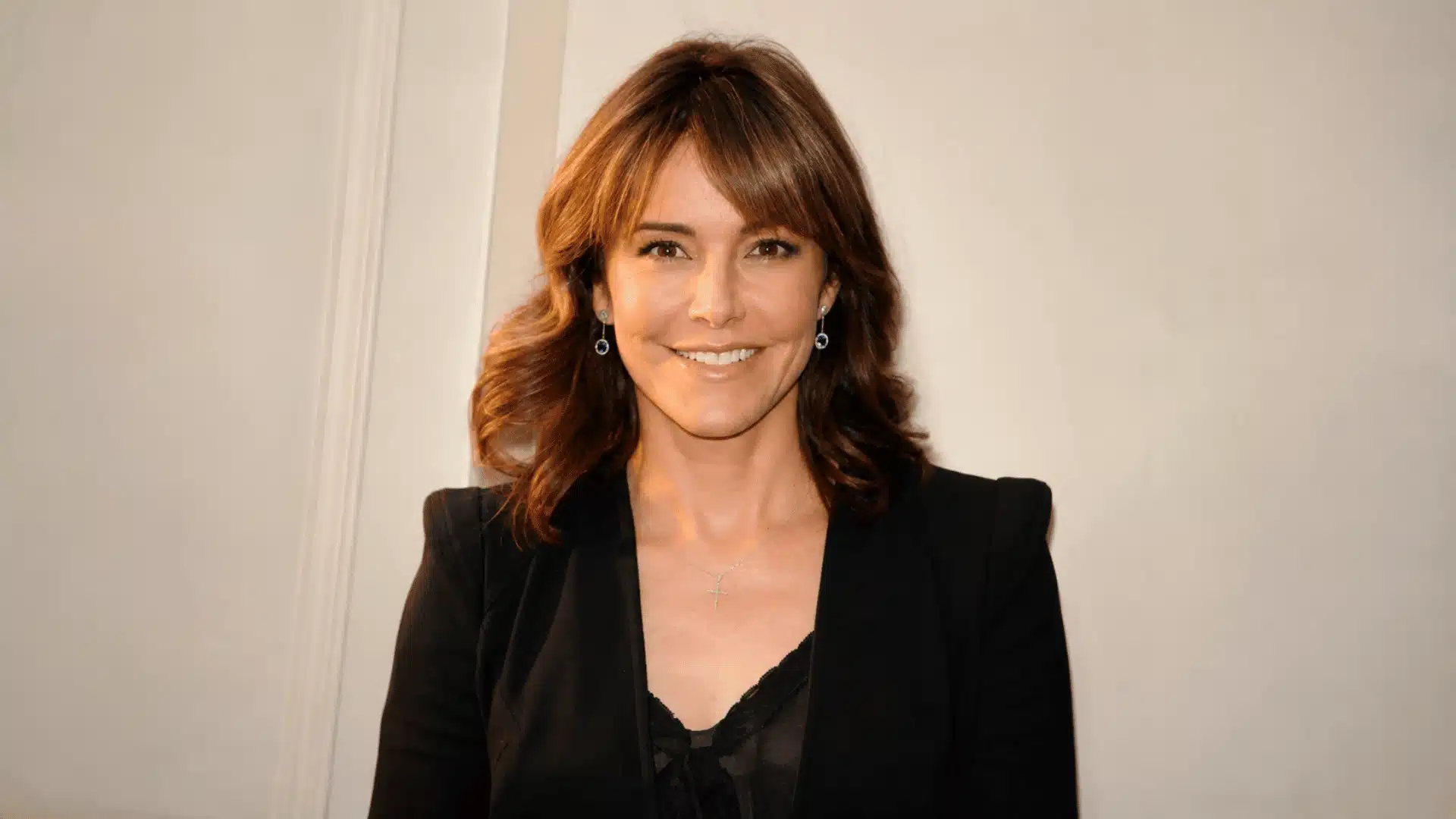 Christa Miller smiling in a black blazer with wavy brown hair, blue earrings, and a cross necklace indoors