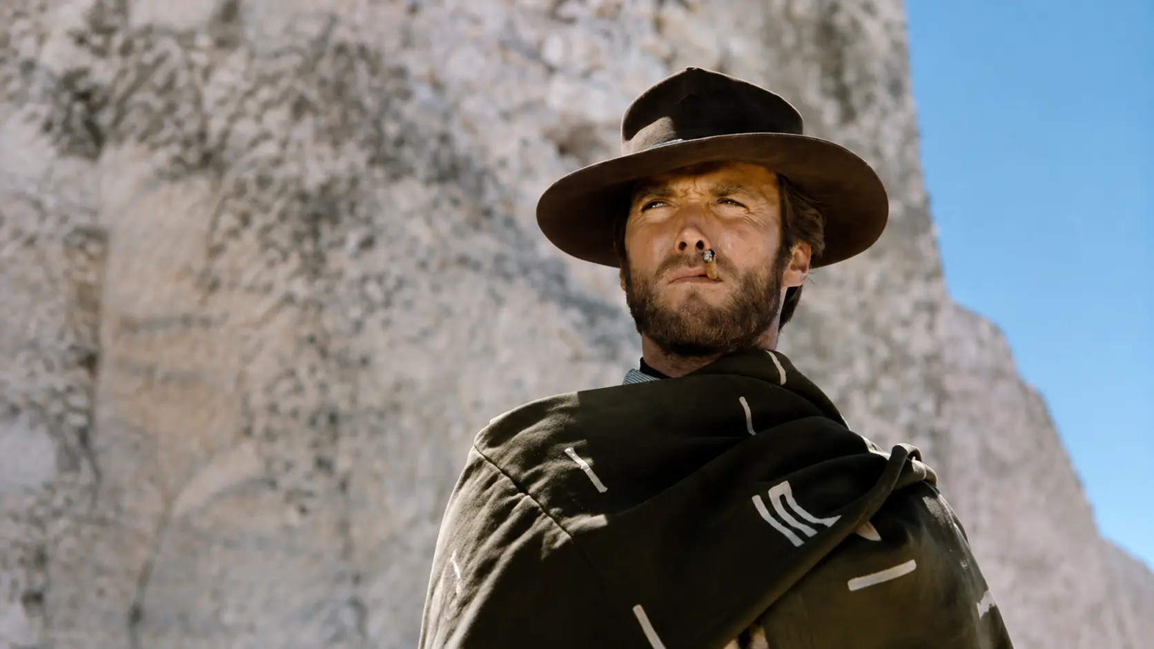 Clint Eastwood wearing a brown hat and dark poncho standing near a rocky landscape under a clear blue sky in a western scene