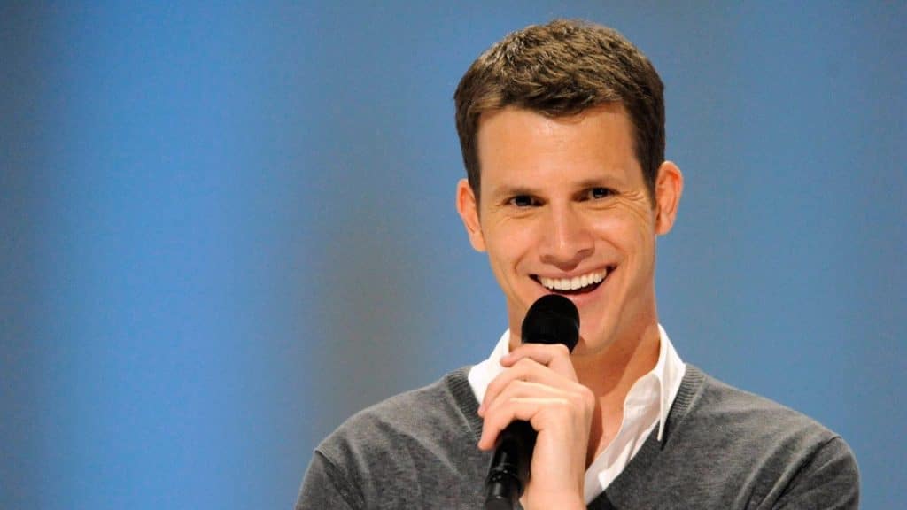 Daniel Tosh smiles while holding a microphone against a blue backdrop for a feature about his net worth and comedy career