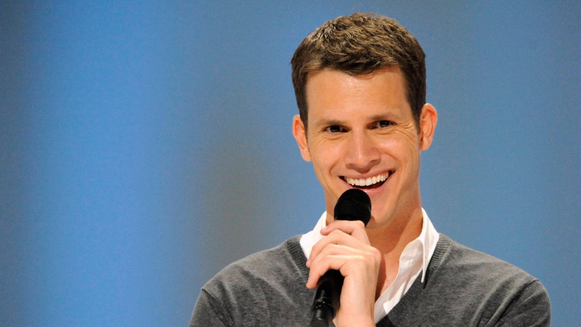 Daniel Tosh smiles while holding a microphone against a blue backdrop for a feature about his net worth and comedy career