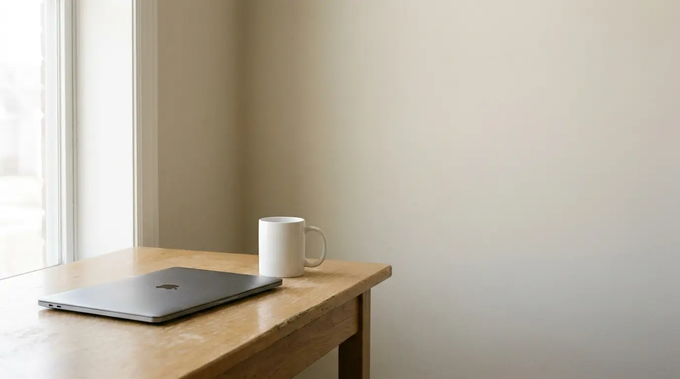 Closed laptop and white mug on wooden desk by sunlit window
