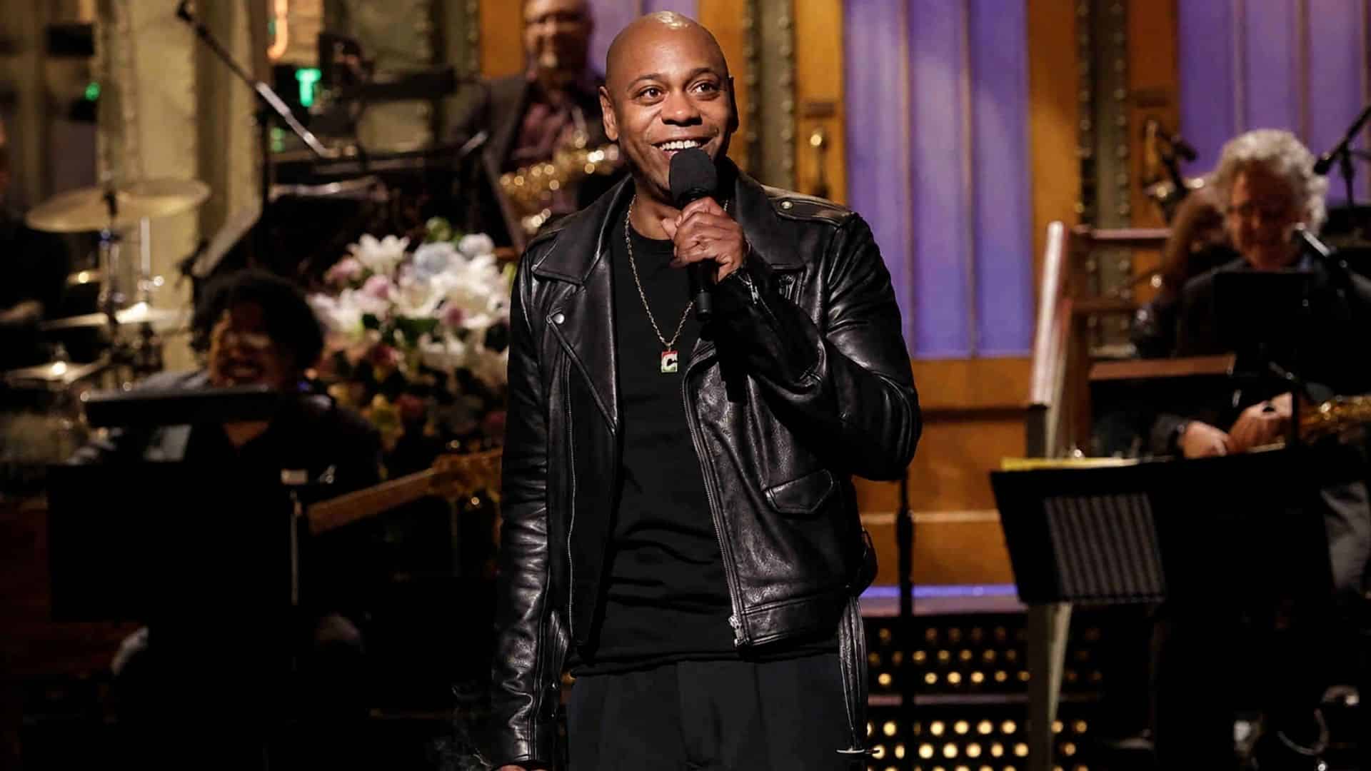 Dave Chappelle smiles while holding a microphone onstage in a black leather jacket with musicians performing behind him