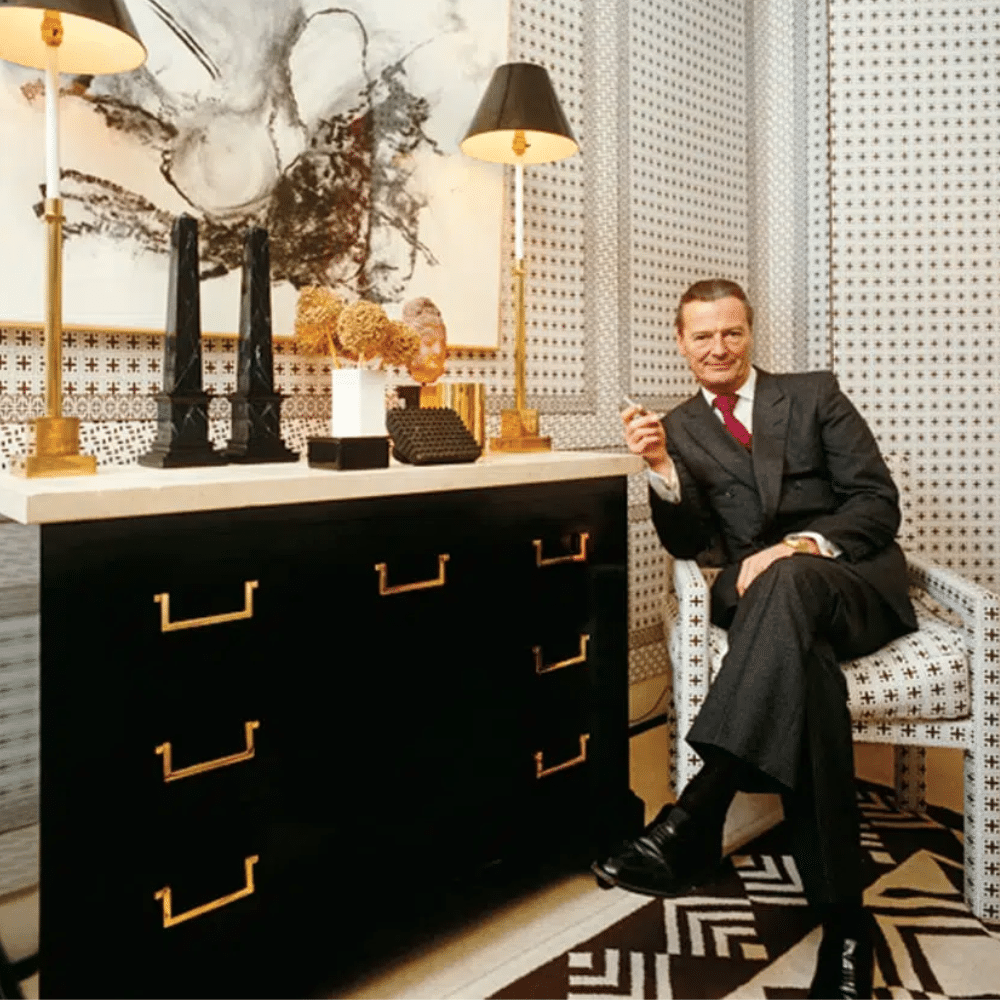 David Hicks in suit seated in patterned room beside black cabinet, table lamps, and abstract artwork.