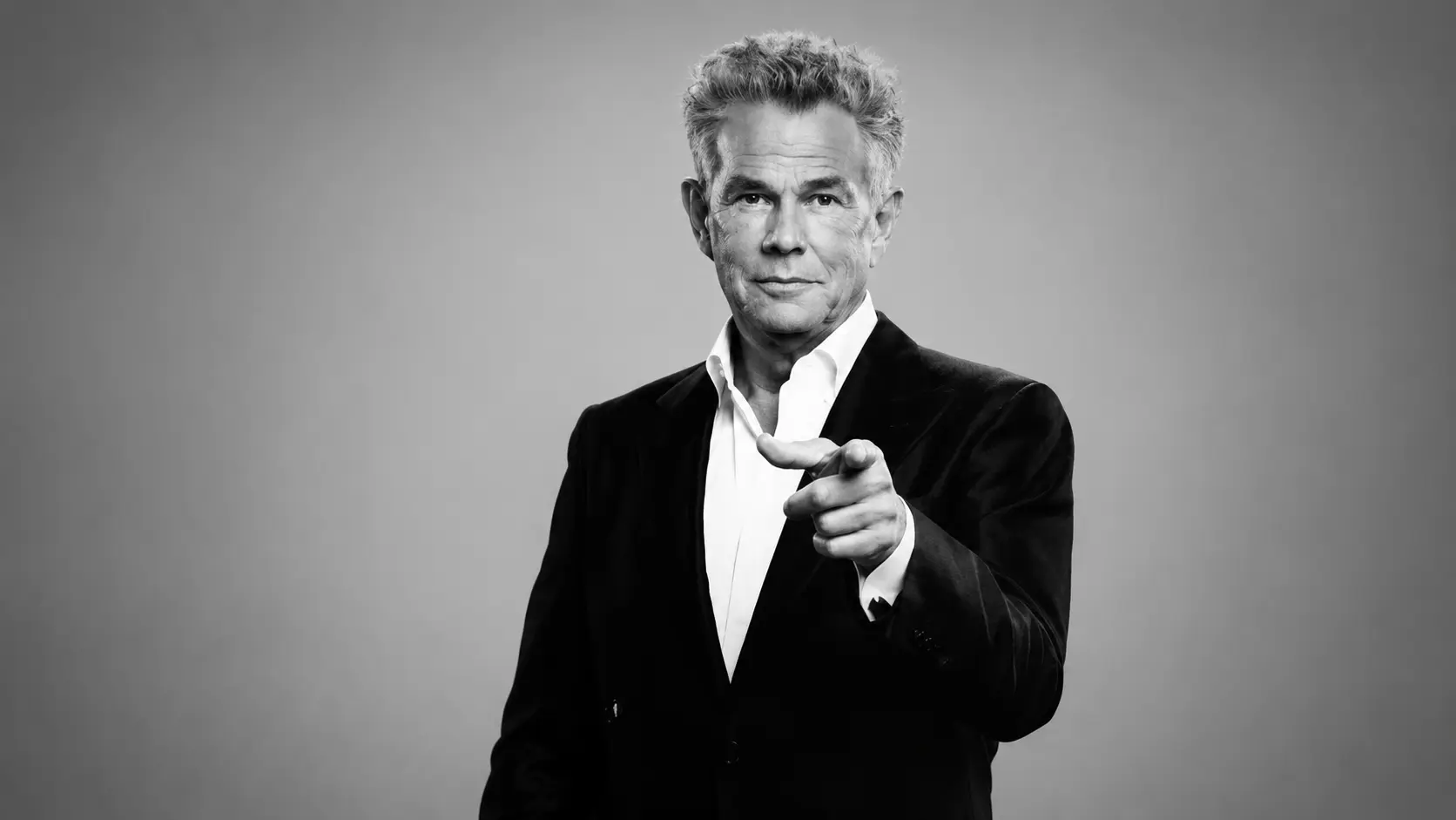 Black and white studio portrait of David Foster in a dark suit pointing toward camera with a soft gray background and lighting