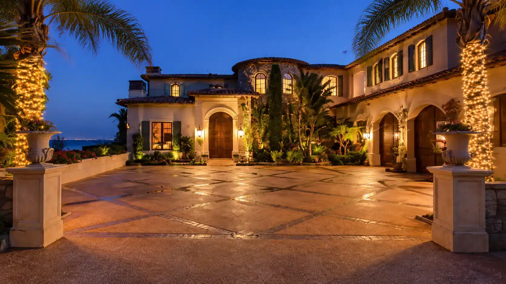 Mediterranean villa exterior at dusk with glowing lights, palm trees, stone courtyard, and arched architecture near the ocean view