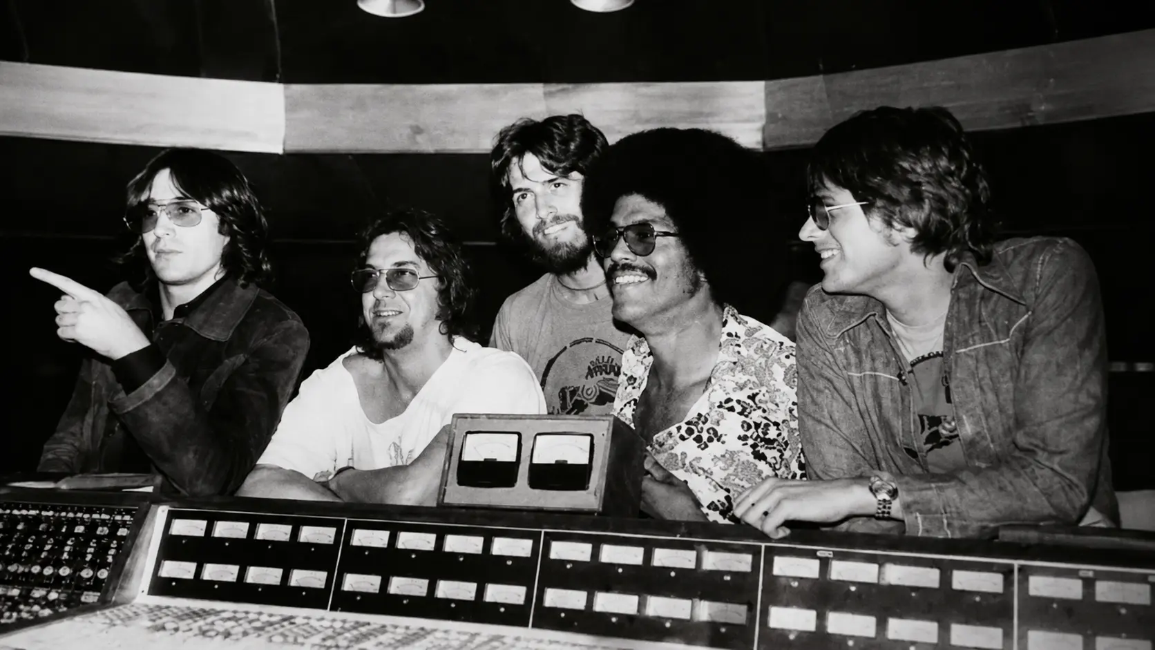 Black and white vintage photo showing musicians gathered in a recording studio behind a large mixing console with equipment