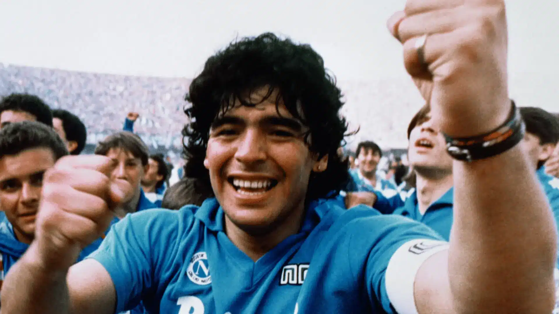 Diego Maradona celebrates with raised fists while wearing a Napoli jersey and captain’s armband.