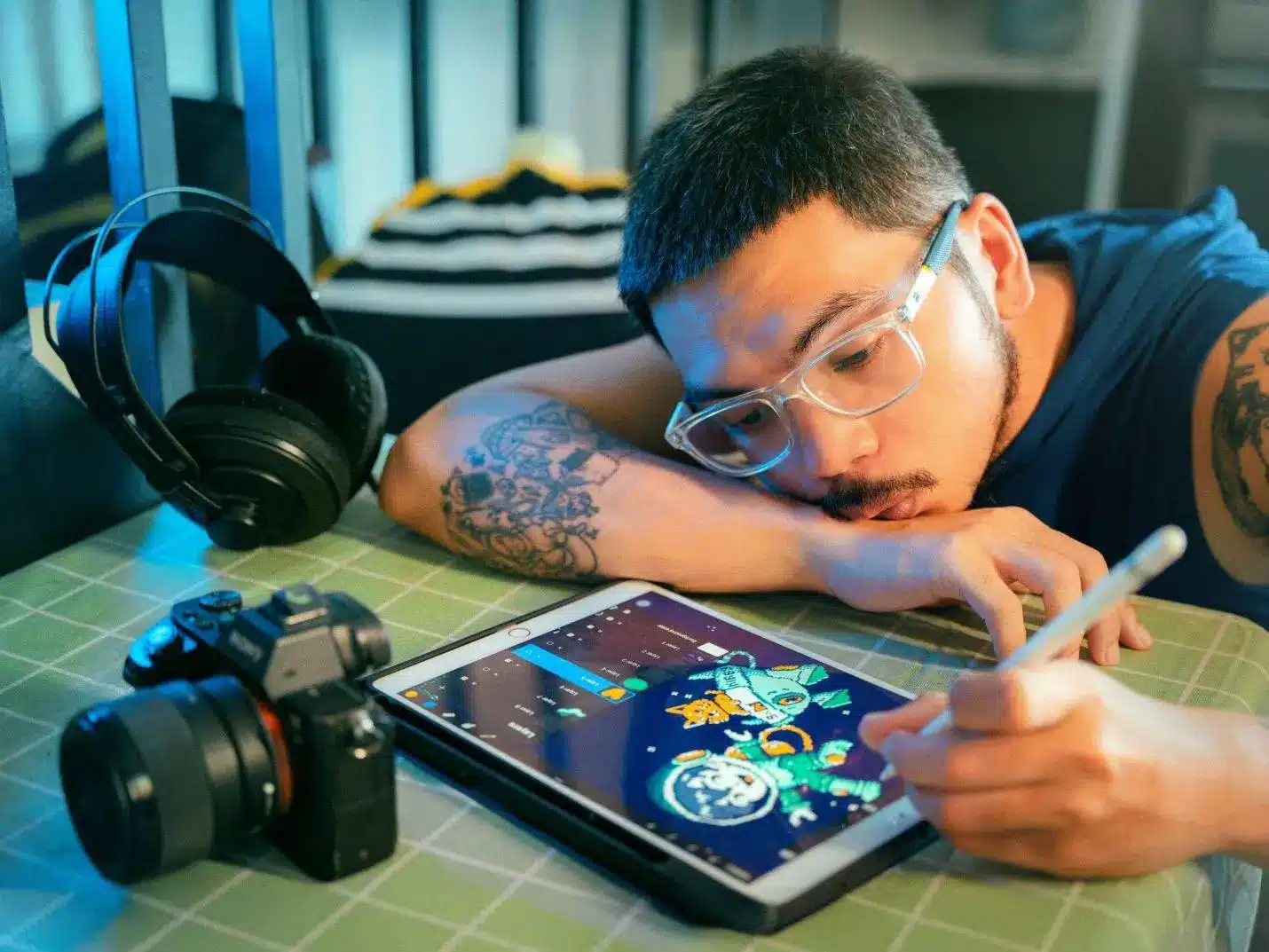 Man with glasses drawing on tablet beside headphones and camera on green table